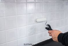 a person using a paint roller on a white tile backsplash with black grout