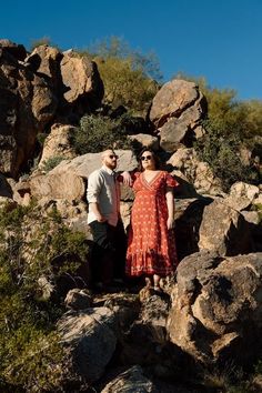 a man and woman standing on top of a rocky hill next to each other with their arms around each other