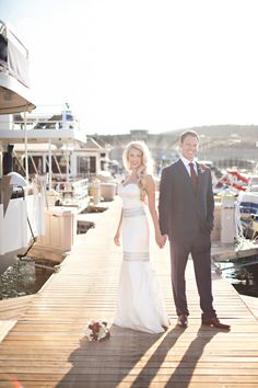 a bride and groom standing on a dock in front of their boats at the marina