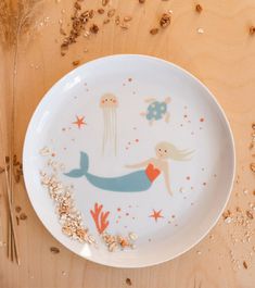 a white plate with an image of a little mermaid on it and seaweed sprinkled around the edge