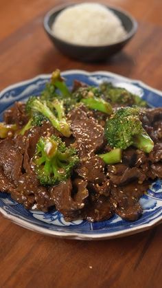 a blue and white plate topped with beef and broccoli next to rice on a wooden table