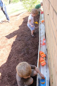 two young boys playing with toy boats in the dirt next to a brick wall and grass