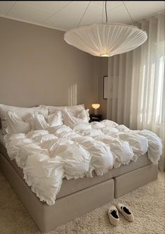 an unmade bed with white sheets and slippers on the floor next to a lamp