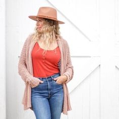 a woman standing in front of a white barn wearing jeans and a red top with a brown hat