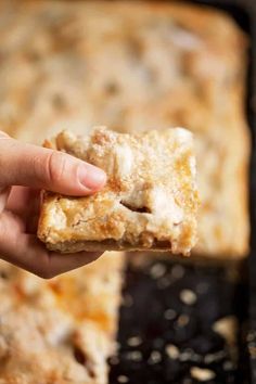 a hand holding up a piece of food in front of a baking pan filled with baked goods