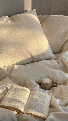 an open book is laying on a bed with white sheets and pillows, next to a cup of coffee