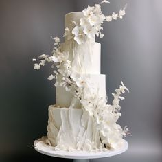 a three tiered cake with white flowers and leaves on the top is displayed in front of a gray background