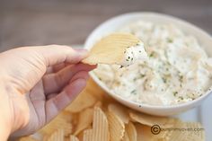 a hand holding a tortilla chip over a bowl of dip and potato chips