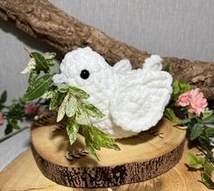 a white crocheted bird sitting on top of a piece of wood next to flowers