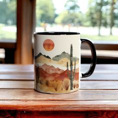 a coffee mug sitting on top of a wooden table with mountains and cactus in the background