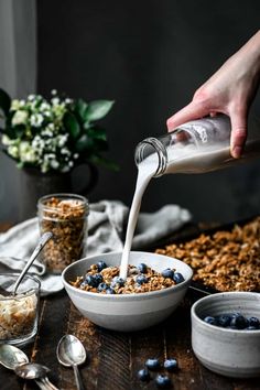 a person pouring milk into a bowl of cereal with blueberries and granola on the side