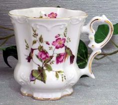 a white vase with pink flowers on it sitting next to a green leafy plant