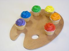 cupcakes with rainbow frosting sitting on a wooden palette holder that is shaped like an artist's palette