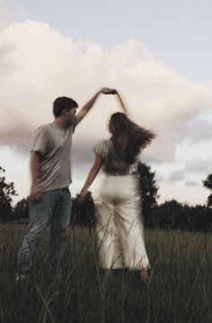 two people standing in tall grass holding hands and looking at the sky with clouds behind them