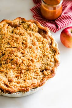 an apple pie sitting on top of a white plate next to apples and a jar of jam