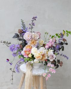 a white vase filled with lots of flowers on top of a wooden stool next to a wall