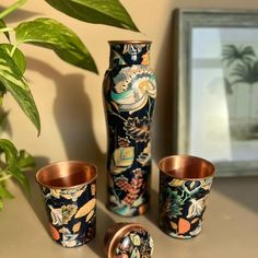 three vases and two cups sitting on a table next to a potted plant