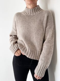 a woman standing against a wall with her hands in her pockets and wearing a turtle neck sweater