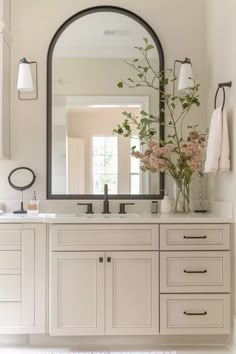 a white bathroom with a large mirror above the sink and flowers in vase on the counter