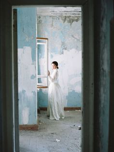 a woman in a white dress standing by a window looking at the wall with peeling paint on it