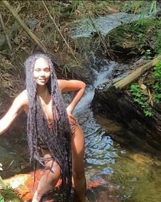 a woman with long hair standing in the water next to a stream and holding onto a rock