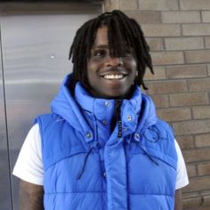 a man with dreadlocks standing in front of a brick wall wearing a blue vest