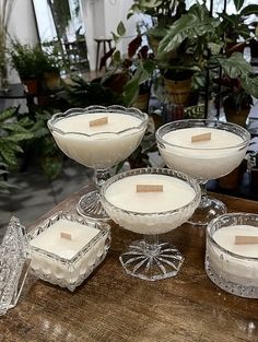 four glasses filled with white liquid sitting on top of a wooden table next to potted plants