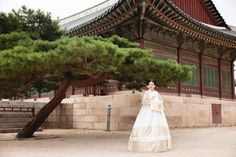 a woman standing in front of a building with a pine tree next to her and wearing a long white dress