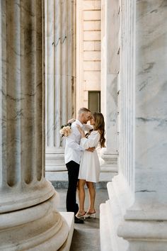 a man and woman standing next to each other in front of columns with their arms around each other