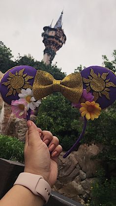 a hand holding up a purple mouse ears with flowers on it and a castle in the background