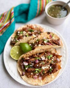 three tacos are sitting on a white plate with limes and cilantro