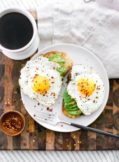 two eggs are on toast with avocado and other toppings next to a cup of coffee