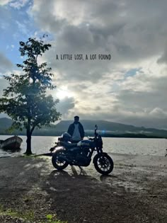 a man sitting on a motorcycle next to a tree near the water with a boat in the background