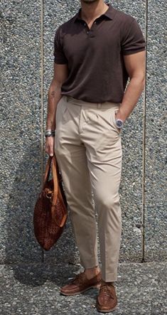 a man standing in front of a wall holding a brown bag and wearing tan pants