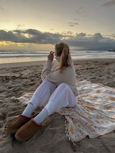 a woman sitting on top of a beach next to the ocean while talking on a cell phone