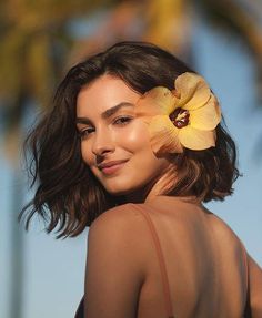 a woman with a yellow flower in her hair and wearing a brown tank top is smiling at the camera