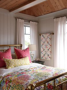 a bed with pink and yellow pillows on top of it next to two windows in a bedroom