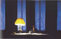 a man sitting at a table in front of a lamp with blue curtains behind him