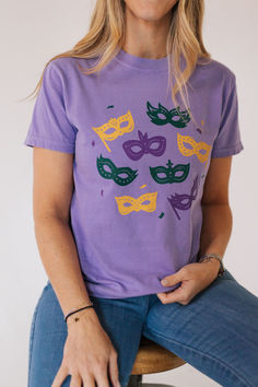 a woman sitting on a stool wearing a purple t - shirt with mardi gras masks