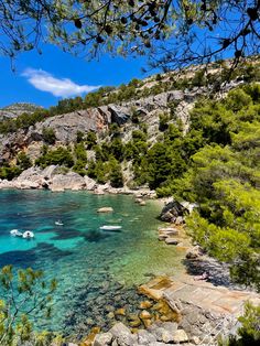 boats floating in the clear blue water near some rocks and trees, on a sunny day