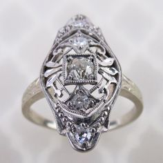 An old European cut diamond is centered on this antique 14k white gold filigree ring. The 3mm .10 carat precious gem is mounted in a square setting. Delicate airy scroll filigree with leaves and millgrain dots decorates the ring. The 13/16 inch long face has a domed gallery with engraved profiles, which is emulated on the ring's shoulders. There are four additional 1.5mm to 1.8mm diamonds set around on the ring. Collectively, this ring has an estimated total weight of .18 cwt diamonds.  The insi Antique Platinum Diamond Ring With Diamond Accents, Victorian Diamond Ring In White Gold, Victorian White Gold Diamond Ring, Victorian Platinum Diamond Ring With Single Cut Diamonds, Victorian Diamond Ring With Diamond Accents, Antique Diamond White Ring With Diamond Accents, Victorian Diamond White Diamond Ring, Victorian Style Diamond Ring With Accents, Victorian Style Diamond White Diamond Ring