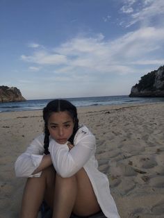 a woman sitting on top of a sandy beach next to the ocean with her arms crossed