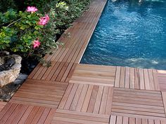 a wooden deck next to a pool surrounded by greenery and pink flowered bushes