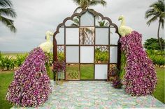 two peacocks are standing in front of a mirror with flowers on the ground and palm trees behind them