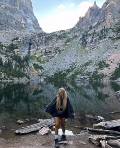 a woman standing in front of a mountain lake with her back turned to the camera