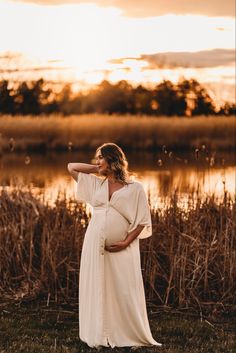 a pregnant woman standing in front of a body of water wearing a white dress and holding her hand on her hip