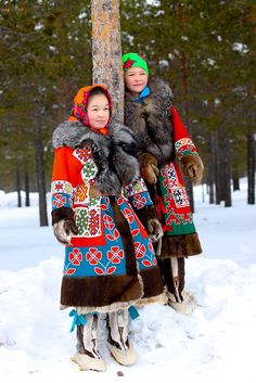 two people in colorful clothing standing next to a tree with snow on the ground and trees behind them