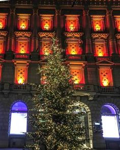 a lit christmas tree in front of a large building with red lights on it's windows