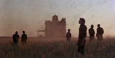 several people standing in a field watching the sun go down on an old farm house