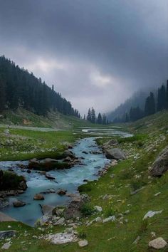 a river running through a lush green hillside covered in rocks and grass under a cloudy sky